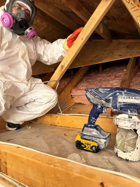 Technician in a Tyvek suit and respirator spraying treatment on attic framing