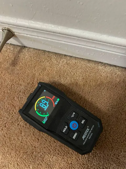 Pinless moisture meter reading 89 on drywall at the baseboard, in the wet range